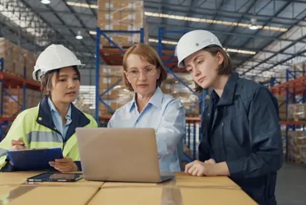 Tres mujeres con casco en almacén revisando información en una laptop