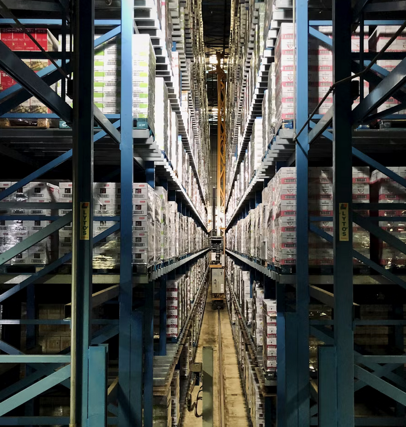 Pasillo de almacén con estanterías altas llenas de pallets organizados, perspectiva desde el centro.