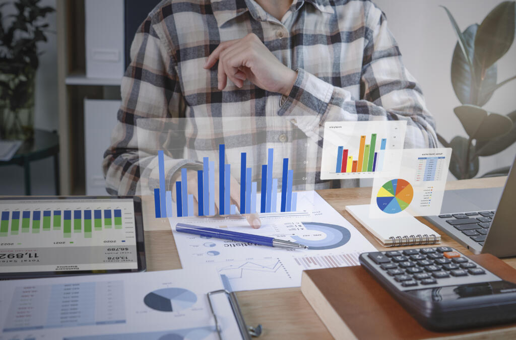 Persona analizando gráficos y estadísticas financieras en una oficina, utilizando tecnología digital y documentos en papel.
