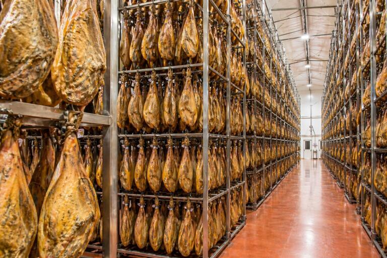 Sala de curado de jamones colgados en estantes metálicos, proceso de producción de jamón ibérico.
